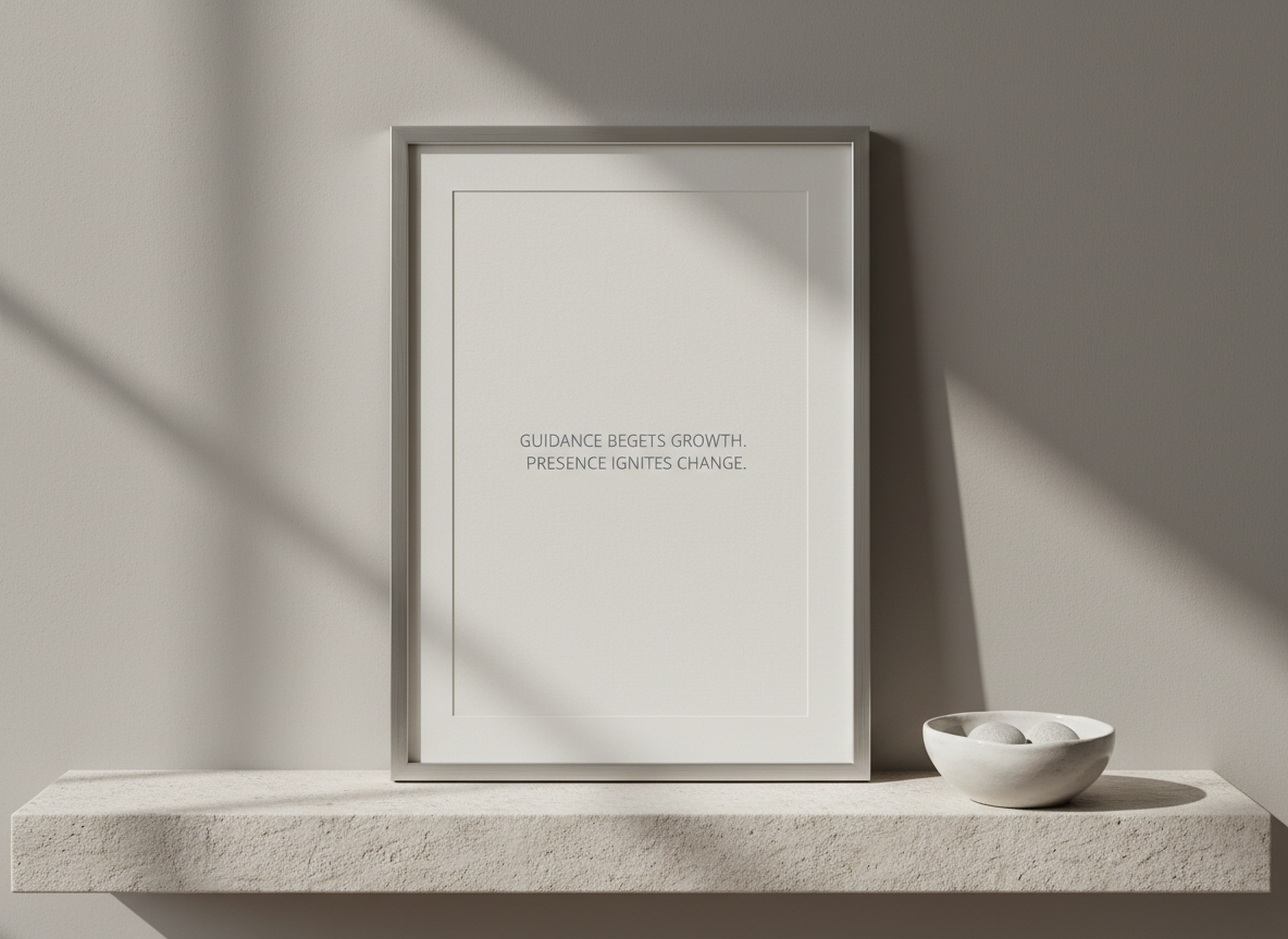 A framed, minimalist statement of therapeutic philosophy displayed on a light stone shelf, the frame in brushed matte aluminum with a narrow border. Inside, on textured off-white paper, understated gray typography reads a short, centered affirmation about guidance and transformation (text not fully legible). Next to the frame, a small, unglazed white ceramic bowl holds three pale gray pebbles. The wall behind is a smooth, warm gray, creating subtle contrast with the shelf. Soft, indirect daylight falls from the left, producing gentle, linear shadows and a quiet glow on the metal frame. Captured straight-on with balanced composition and ample negative space, the image feels stable, thoughtful, and corporate-professional in photographic realism.